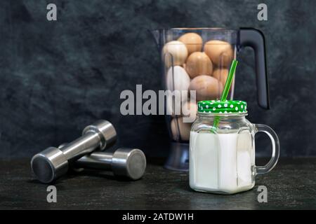 Secouez les protéines dans un mug à l'arrière-plan d'un bol mélangeur avec des œufs de poulet et des haltères sur un fond sombre. Contexte. Concept de nutriti sportif Banque D'Images