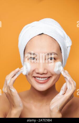 Une jeune femme asiatique heureuse qui applique de la crème pour le visage tout en portant une serviette et en touchant son visage. Isolé sur fond orange Banque D'Images