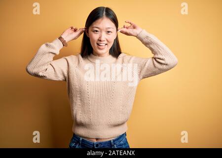 Jeune femme asiatique portant un pull décontracté sur fond jaune isolé se moquer des oreilles avec les doigts, geste drôle. Problème d'audition Banque D'Images