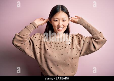 Jeune femme asiatique belle portant la mode et élégant pull sur fond solé rose se laçant les oreilles avec les doigts, geste drôle. Auditio Banque D'Images