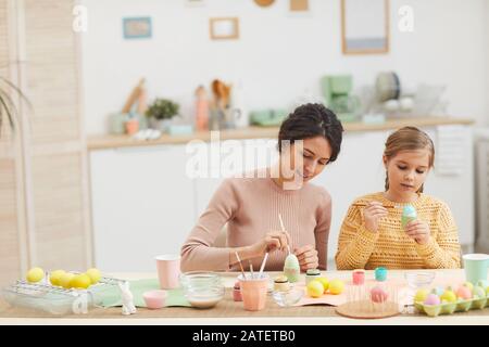 Portrait grand angle de la mère et de la fille peindre des oeufs de Pâques couleur pastel assis à la table dans la cuisine intérieure confortable, espace de copie Banque D'Images