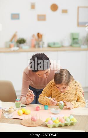 Portrait vertical de la mère et de la fille peignant des œufs de Pâques ensemble assis à la table dans une cuisine intérieure confortable, espace copie Banque D'Images