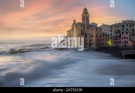 Église de Camogli, Ligurie, Italie au coucher du soleil contre un ciel jaune coloré avec des vagues se brisant sur la plage au premier plan Banque D'Images
