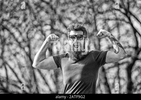 L'homme barbu montre des muscles, des biceps le jour ensoleillé. Concept de masculinité. L'homme avec une barbe longue a l'air énergique et gai. Homme avec barbe et moustache sur le visage heureux, branches sur le fond, démoqué. Banque D'Images