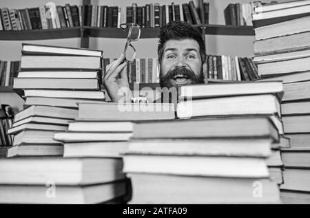 Homme sur le visage heureux entre des piles de livres dans la bibliothèque, des étagères sur fond. Professeur ou étudiant avec barbe porte des lunettes, assis à la table avec des livres, démoqué. Concept de recherche scientifique. Banque D'Images