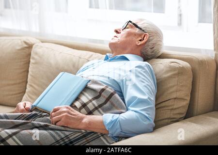 Homme senior fatigué couvert avec des aides au rodage sur le canapé avec livre ouvert Banque D'Images