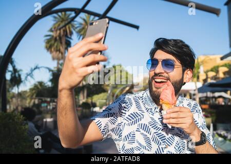 Portrait de jeune homme profitant du temps ensoleillé, prendre un selfie avec le téléphone et manger une glace à l'extérieur. Banque D'Images