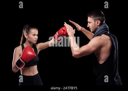 Jeune femme sérieuse dans l'activewear et gants de boxe frapper l'entraîneur Banque D'Images