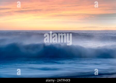 Brisant les vagues avec un jet brumeux au coucher du soleil sur la mer sous un ciel orange coloré Banque D'Images