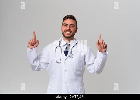 Beau moderne docteur blanc robe médicale dans studio gris de la publicité de fond de votre produit regardant l'appareil photo et pointant le doigt vers le positiv Banque D'Images