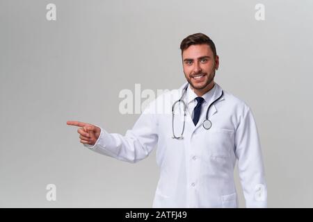 Beau moderne docteur blanc robe médicale dans studio gris de la publicité de fond de votre produit regardant l'appareil photo et pointant le doigt vers le positiv Banque D'Images