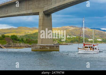 Pont de l'île de Skye avec bateau de croisière blanc et rouge passant sous le célèbre pont. Montagnes et cottages en arrière-plan. Paysage. Banque D'Images