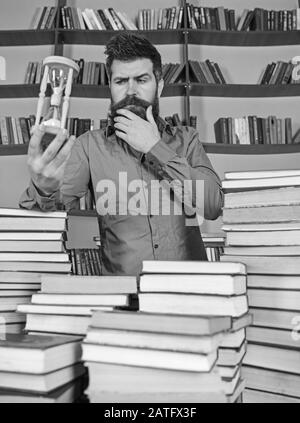 Concept de bibliothécaire. L'homme sur le visage réfléchi se tient entre des piles de livres, tout en tient sablier, des étagères à livres sur l'arrière-plan. Professeur, scientifique avec des barbes à la table avec des livres, défocused. Banque D'Images