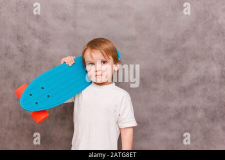 Petit garçon portant une tenue décontractée, tenez son skateboard sur son épaule, debout sur un fond gris. Vente concept équipement sportif Banque D'Images