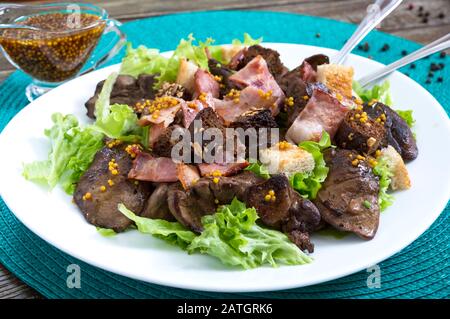 Salade chaude et saine de foie de poulet, de croutons de seigle, de bacon fumé, de salade verte et de sauce moutarde dans une assiette blanche sur fond de bois. Gros plan Banque D'Images