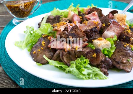 Salade chaude et saine de foie de poulet, de croutons de seigle, de bacon fumé, de salade verte et de sauce moutarde dans une assiette blanche sur fond de bois. Gros plan Banque D'Images