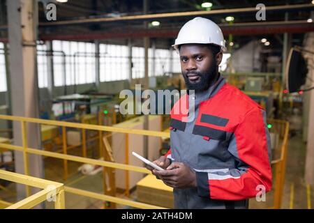 Portrait d'un homme noir barbu confiant dans un casque de protection à l'aide d'une tablette numérique en usine Banque D'Images