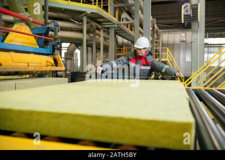 Travailleur de la chaîne de production dans un casque résistant qui se tient sur le tapis du convoyeur et mesurant les matériaux de construction à l'usine Banque D'Images