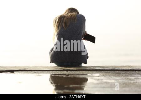 Vue arrière portrait d'une fille triste tenant le téléphone mobile se plaignant assis sur une corniche Banque D'Images