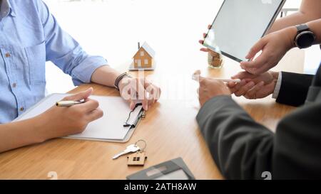 Vue latérale de l'agent immobilier présentant au client une tablette à écran vide, un modèle de maison et une calculatrice sur un bureau en bois Banque D'Images