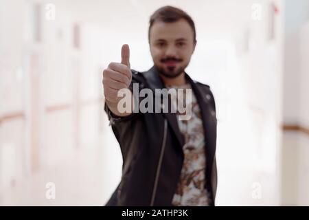 Portrait d'un homme souriant et barbu se faisant un geste de pouce dans un couloir ou une université de hall. Approbation du concept, recommander, comme un nouveau produit. Banque D'Images