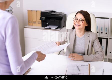 Vue sur l'épaule de la dame senior donnant forme remplie au sourire sympathique travailleur social au bureau Banque D'Images