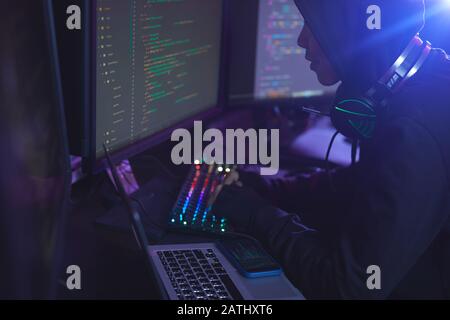 Vue latérale sur un pirate informatique non reconnaissable portant une capuche lors de la programmation dans une pièce sombre, espace de copie Banque D'Images
