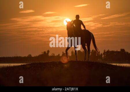Silhouette jeune homme à cheval au coucher du soleil sur la colline au bord du lac Banque D'Images