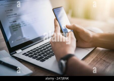 L'homme travaille à la maison et utilise un smartphone et un ordinateur portable. Silhouette d'un homme occupé. Une main tient et utilise le téléphone, l'autre écrit dessus Banque D'Images