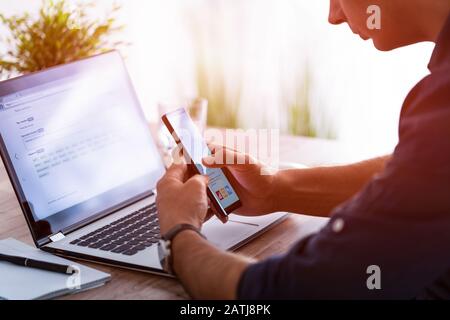 L'homme travaille à la maison et utilise un smartphone et un ordinateur portable. Silhouette d'un homme occupé. Une main tient et utilise le téléphone, l'autre écrit dessus Banque D'Images