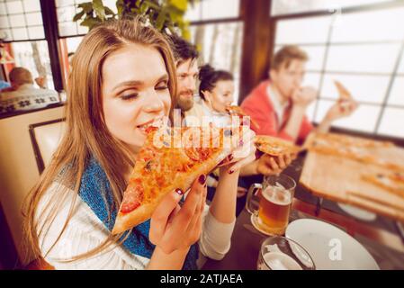 Une jeune fille morde un grand morceau de pizza qui l'a calquée les yeux dans le plaisir. Des amis se fêtes au pub. Gros plan portrait Banque D'Images