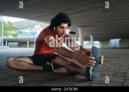 Jeune coureur de sexe masculin écoutant de la musique sur l'oreillette qui s'étire sur ses jambes avant de courir et de faire de l'exercice dans la rue sous le pont Banque D'Images