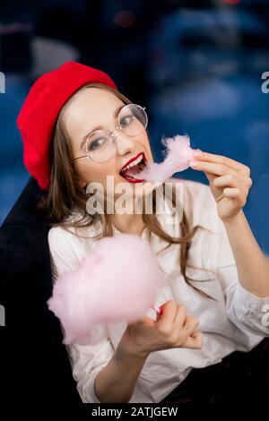 Jeune femme en béret rouge et verres a un grand dessert sucré avec des bonbons en coton rose dans un café en France Banque D'Images