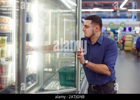 Homme dans un supermarché devant le congélateur à la recherche de ses aliments congelés préférés. Banque D'Images