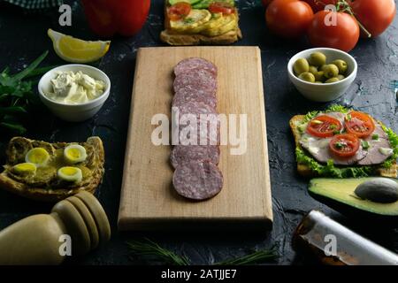 Salami en tranches sur une planche en bois, autour de la planche des olives avec d'autres sandwichs, tomates, citron et sauce. Banque D'Images