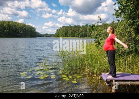 Une jeune femme enceinte pond sur une jetée en bois sur un lac forestier pendant une chaude journée ensoleillée se répandant les mains en profitant de la nature - grossesse saine Banque D'Images