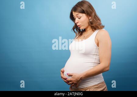 Jeune femme enceinte portant une chemise blanche tenant son ventre ayant des contractions s'occuper de son bébé à naître. Studio tourné sur fond bleu Banque D'Images