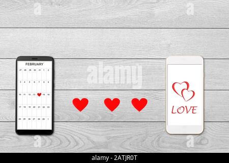 Concept de Saint-Valentin, sur fond de bois blanc isolé coeurs de papier rouge et deux smartphones, calendrier et accueil sur l'écran du smartphone Banque D'Images