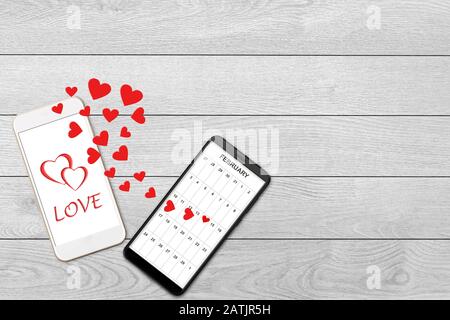 Concept de Saint-Valentin, message d'amour - coeurs sortant de deux smartphones, isolés sur fond blanc en bois, calendrier et accueil sur smartphone s. Banque D'Images