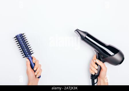 Les mains des femmes tenant le sèche-cheveux et la brosse à cheveux sur fond blanc. Composition de pose plate minimale des outils pour le coiffage des cheveux. Banque D'Images