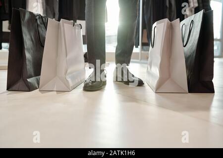 Gros plan d'un homme non reconnaissable dans des chaussures formelles se tenant entre des sacs de shopping complets de vêtements dans le magasin pour hommes Banque D'Images