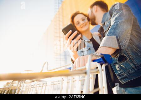 Un jeune couple choisit un magasin par téléphone, l'homme tient un chariot de chariot de supermarché Banque D'Images