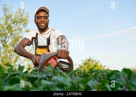 Jardinier professionnel africain masculin en uniforme spécial et lunettes prenant soin des buissons verts avec taille-haie à l'extérieur. Homme souriant utilisant les technologies modernes au travail Banque D'Images