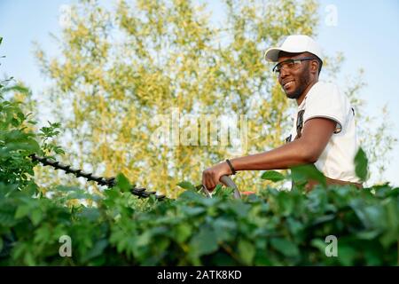Jardinier masculin africain portant des combinaisons avec des lunettes de protection travaillant avec des équipements de jardin professionnels. Heureux jeune homme tailler des buissons. Concept de travail saisonnier Banque D'Images