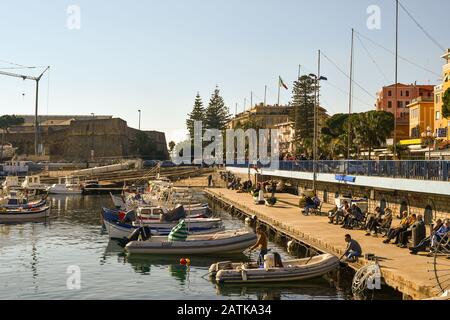 Vue sur le Vieux-Port avec des gens et des touristes marchant sur la promenade et profitant du soleil assis sur les bancs du quai, Sanremo, Ligurie Italie Banque D'Images