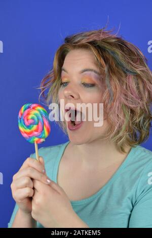 portrait d'une jeune femme blonde avec des cheveux colorés regardant heureux d'une grande lollipop dans ses mains et de penser à s'y mordre Banque D'Images