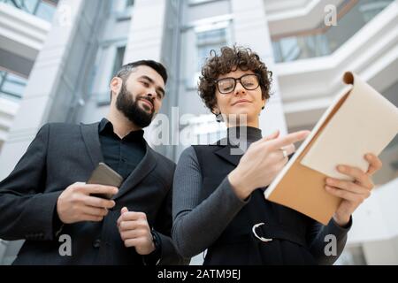 Femme d'affaires aux cheveux bouclés dans des lunettes pointant dans le presse-papiers tout en expliquant la stratégie de développement à un collègue Banque D'Images