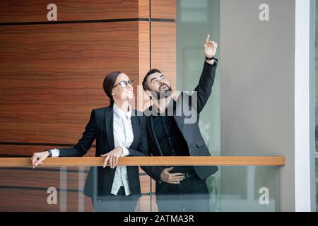 Jeune Manager barbu en veste debout sur le balcon du centre de bureau et pointant vers le haut tout en racontant à un collègue l'expansion de l'entreprise Banque D'Images