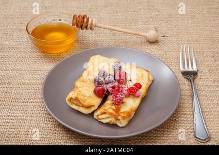 Crêpes maison remplies de fromage cottage et surmontées de framboises et de mûres surgelées sur plaque, fourchette et miel dans un bol en verre sur toile de rackCloth. Banque D'Images