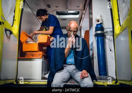L'homme souffle à travers le masque à oxygène, la jeune infirmière en uniforme médical ouvre le sac médical dans la voiture d'ambulance Banque D'Images